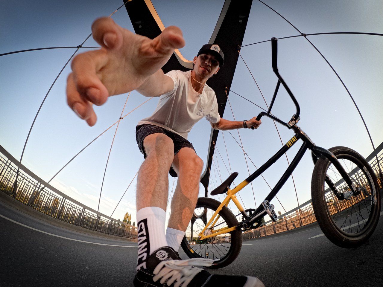 Robin Meza auf einer Brücke neben seinem BMX-Rad, mit Fischaugenobjektiv von unten fotografiert, eine Hand zur Kamera ausgestreckt.
