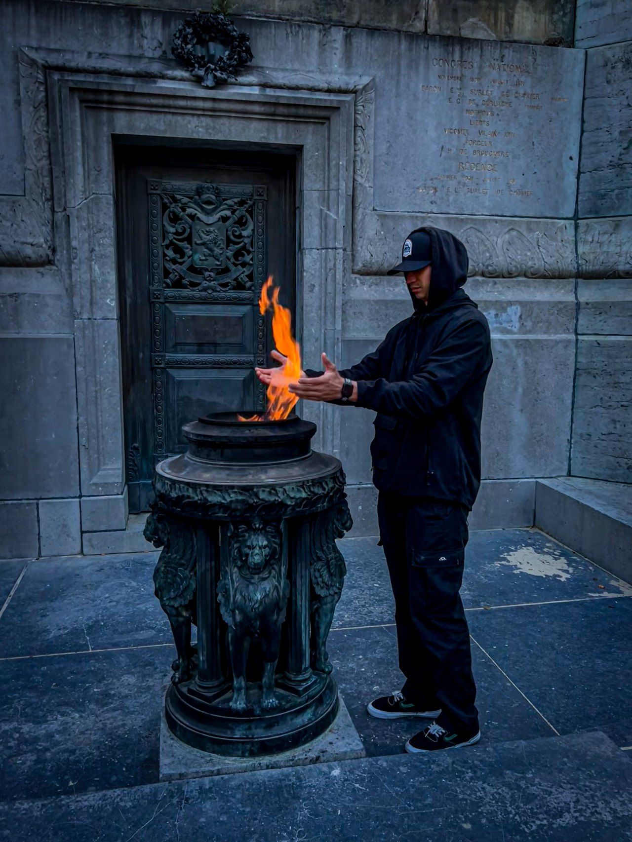 Robin Meza in schwarzem Kapuzenpullover und Cap wärmt sich die Hände an einer Flamme vor einem steinernen Denkmal.