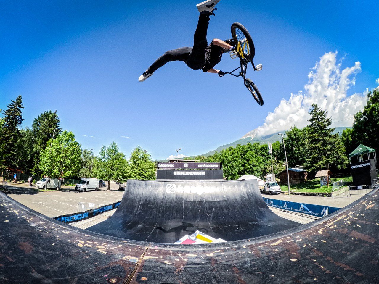 Robin Meza kopfüber in der Luft über einer schwarzen Holzrampe, das BMX-Rad über sich, dahinter blauer Himmel, Bäume und Berge.