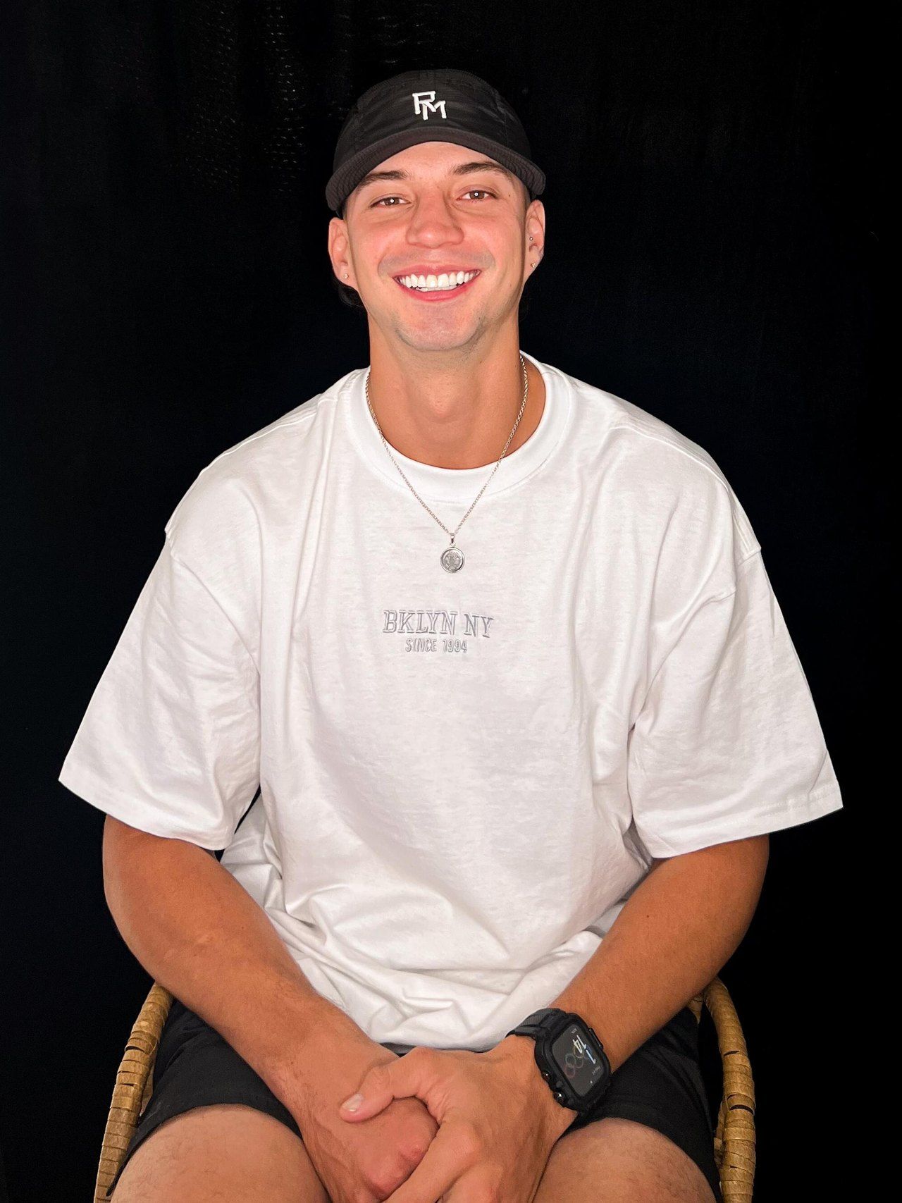 Porträt von Robin Meza, lachend, in weißem T-Shirt und schwarzer Cap vor dunklem Hintergrund.