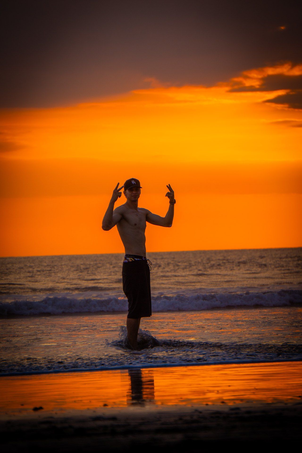 Robin Meza steht bei Sonnenuntergang im flachen Wasser am Strand und zeigt mit beiden Händen das Peace-Zeichen.